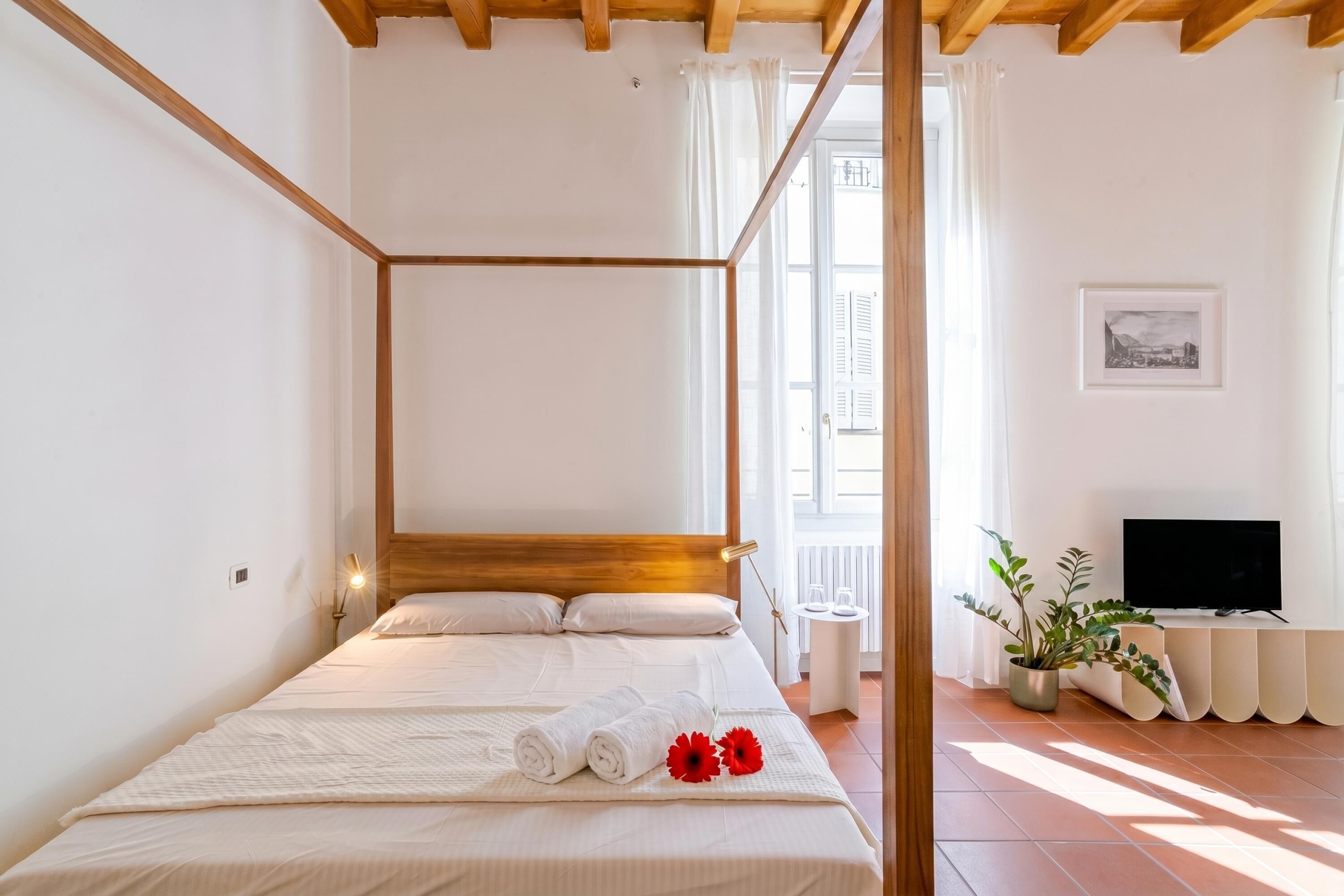 Une chambre à coucher avec un lit à baldaquin, une télévision, une plante en pot et une fenêtre avec des rideaux.