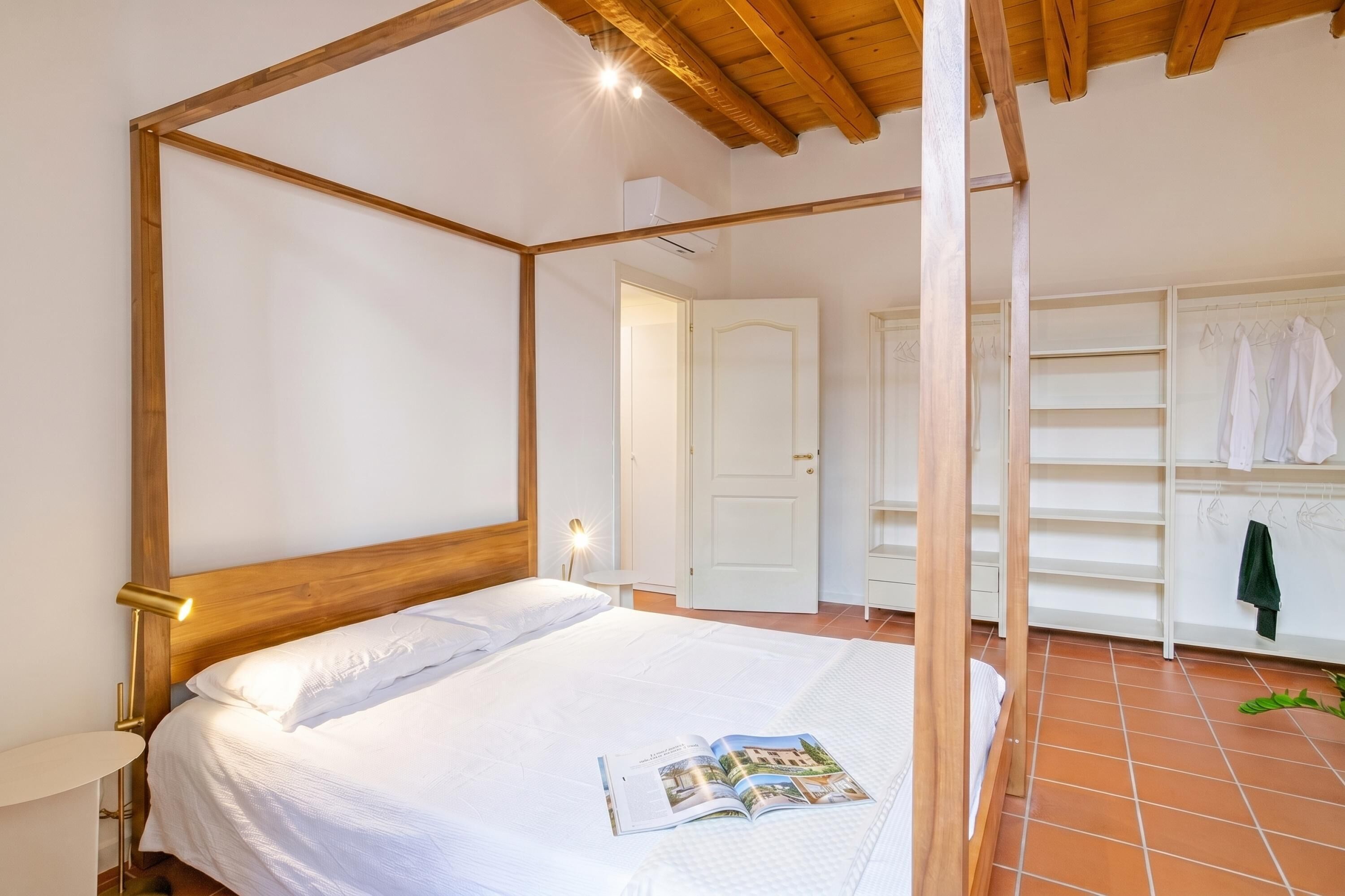 Une chambre avec un lit à baldaquin, une table de chevet, une lampe de lecture, un magazine sur le lit, une porte blanche et un portant à vêtements avec des vêtements suspendus.
