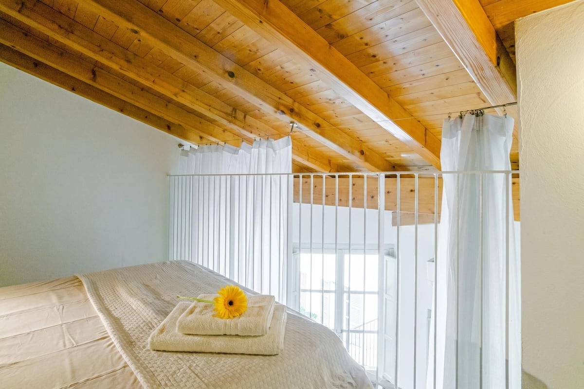 Une chambre avec un plafond en bois, un lit recouvert d’une couette blanche, un rideau blanc et une fenêtre donnant sur un paysage agréable.