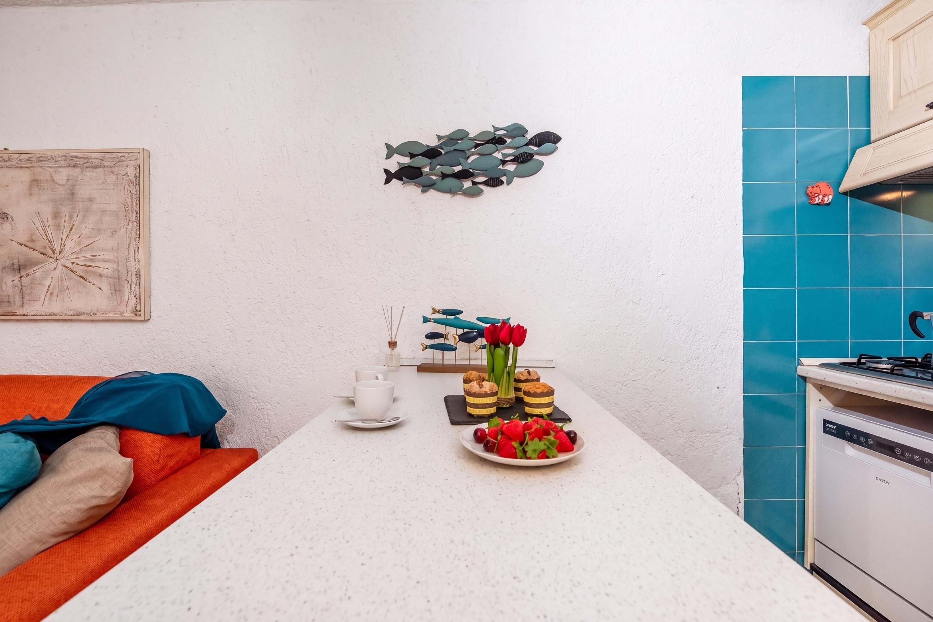 Eine Küche mit weißer Arbeitsplatte, einer blauen Fliesenwand, Wandkunst mit einem Fischmotiv, ein kleines Flugzeug-Dekorationsobjekt und ein Teller mit Erdbeeren und Kirschen.