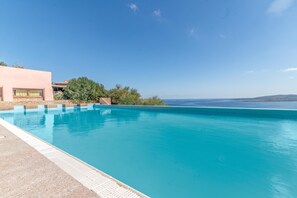 A clear blue swimming pool with a stone wall and a building with a red roof in the background. - Ladunia 2 (Olbia)