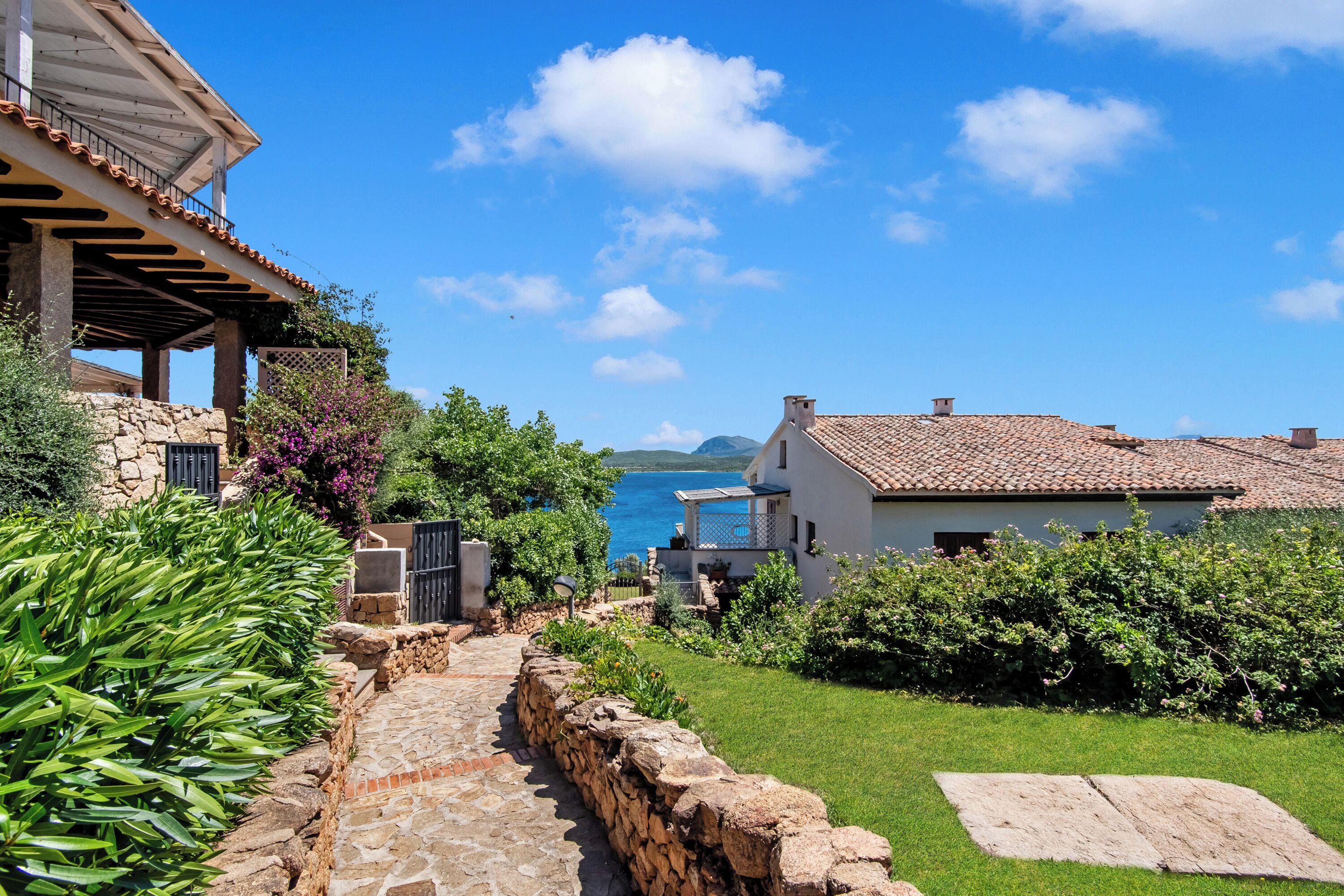 Um imóvel costeiro com um caminho de pedra que leva a um edifício branco com telhado de telhas, rodeado por vegetação exuberante e vista para o mar.