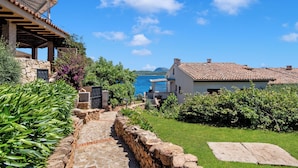 A coastal property with a stone pathway leading to a white building with a tiled roof, surrounded by lush greenery and a view of the sea. - Ladunia 2 (Olbia)
