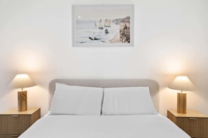 A bedroom with a bed, two bedside tables with lamps, a nightstand, and a framed picture of a coastal scene. - Bright 1-Bed Stay Near Crown Melbourne (South Melbourne)