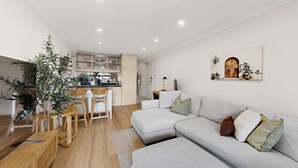 En moderne stue med en grå hjørnesofa, et spiseområde med et træbord og stole, samt et køkken med hvide skabe.