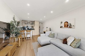 A modern living room with a grey sectional sofa, a dining area with a wooden table and chairs, and a kitchen with white cabinets. - Bright 1-Bed Apartment in Neutral Bay (Neutral Bay)