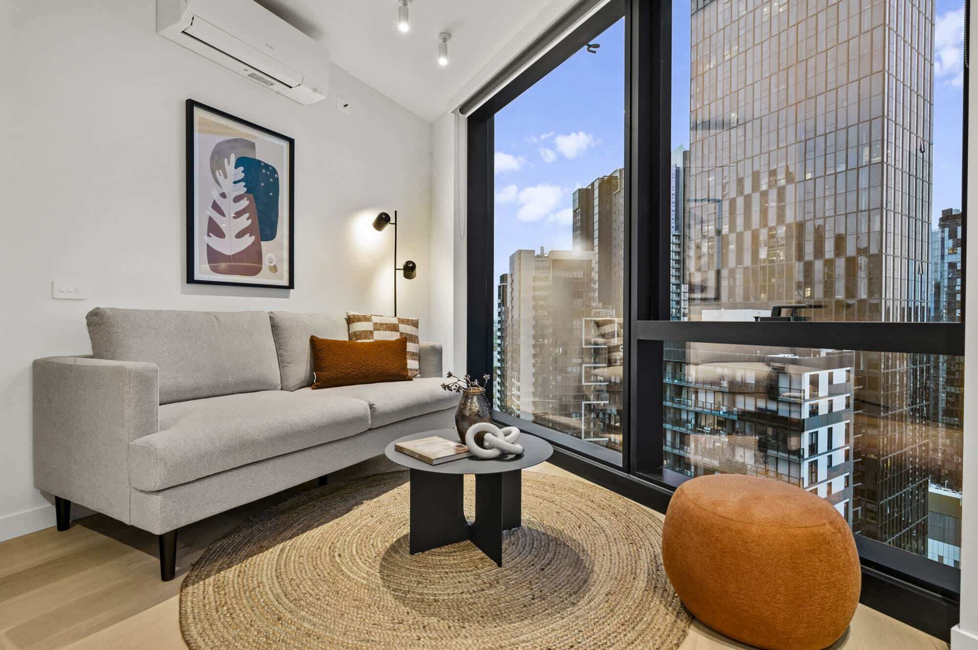 A modern living room with a grey sofa, a round coffee table, and a large window offering a city view.