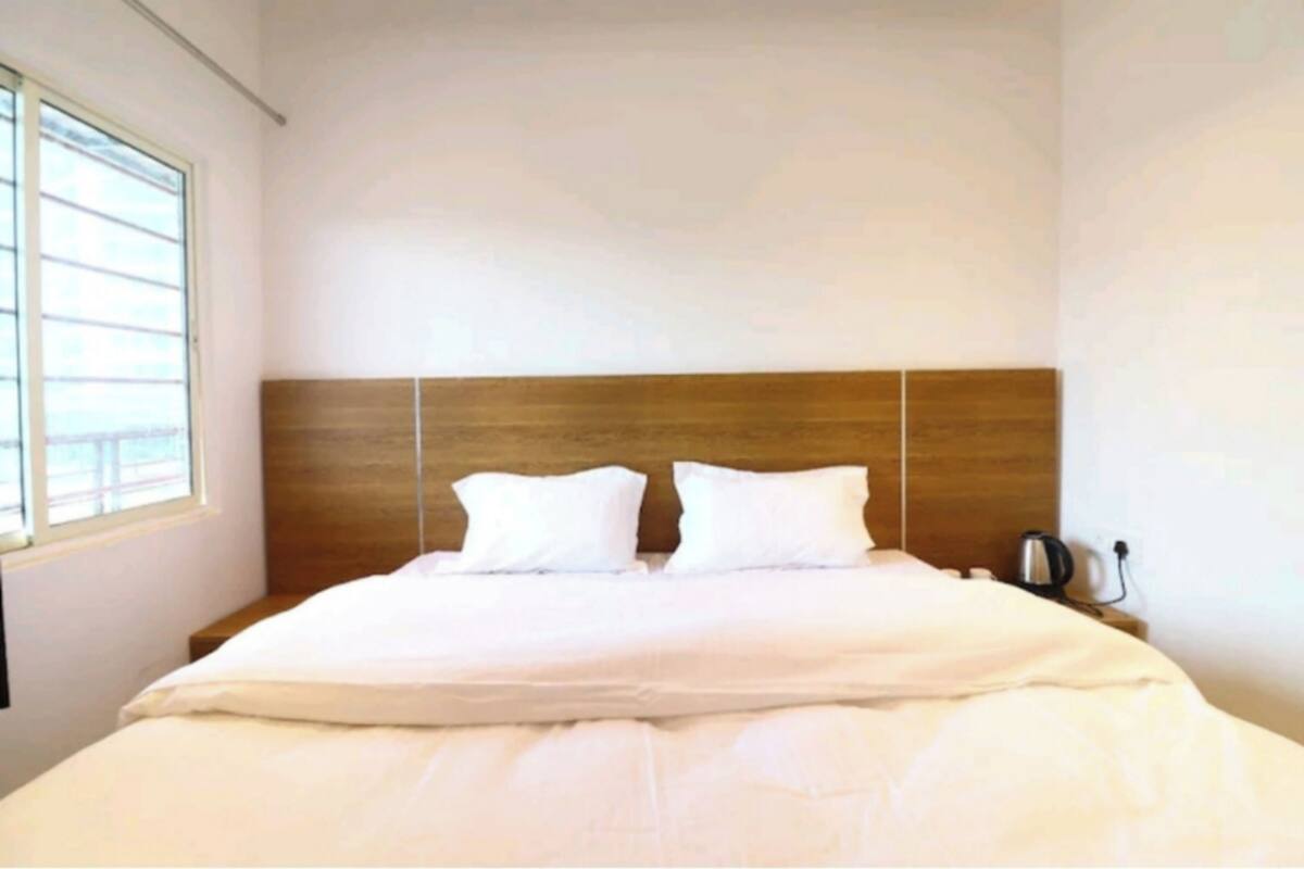 A hotel room with a large bed, a wooden headboard, a bedside table with a lamp, and a window.
