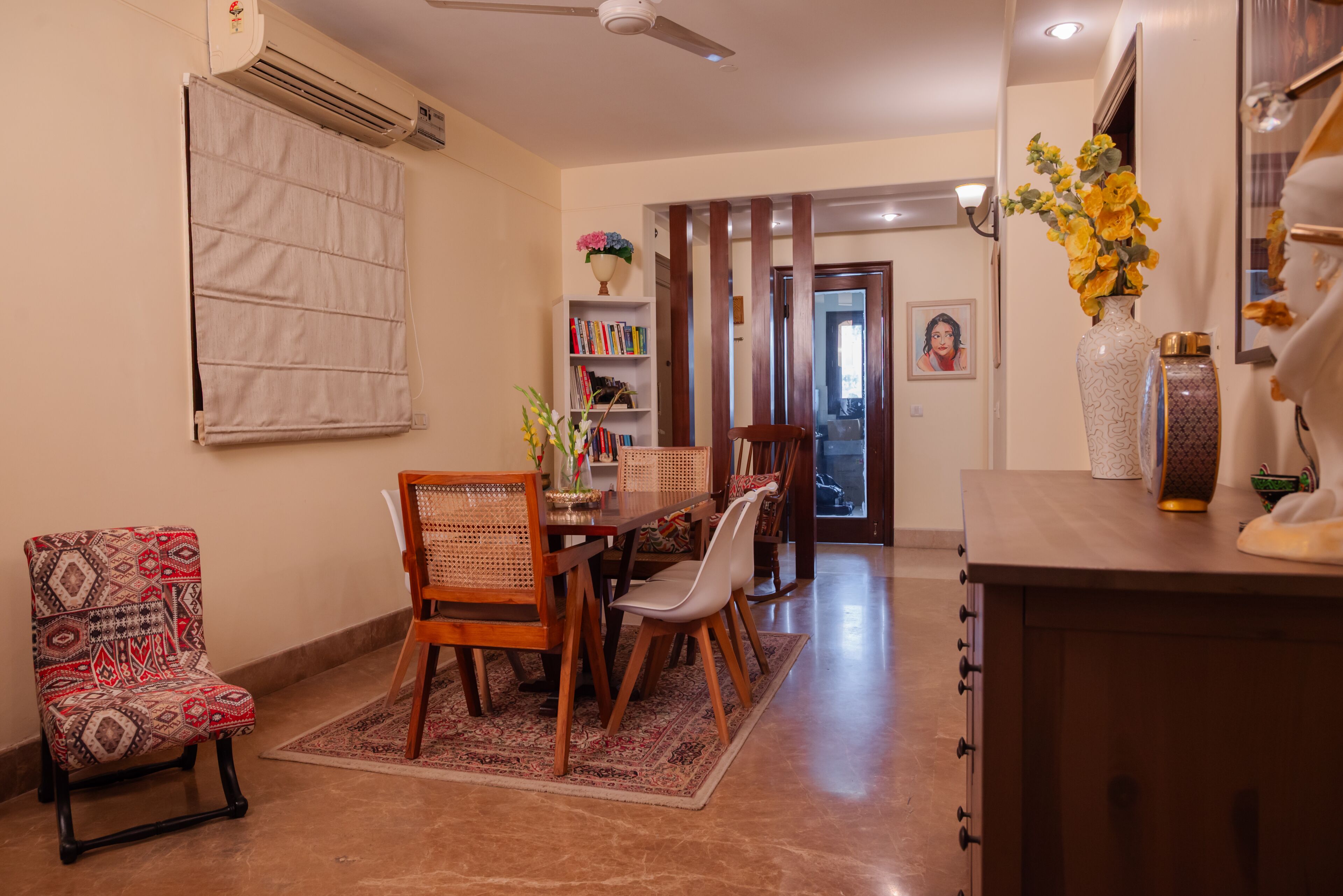 A dining area with a wooden table, chairs, a patterned armchair, a bookshelf, and a vase with flowers.