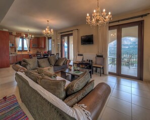 A spacious living room with a sofa, a dining area, a TV, and a balcony with a view. - Hidden Gem in Crete — Totally Private Villa for up to 9 Guests (Rethymno)