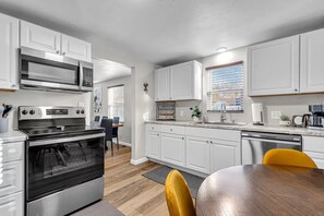 A modern kitchen with white cabinetry, a built-in oven, a microwave, and a dining area with a wooden table and yellow chairs. - Victory House. Close to Lambeau Field! (Green Bay)