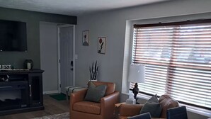 Un saló amb un sofà de cuir marró, una taula de dinar de fusta amb cadires, una televisió i una finestra amb persianes.