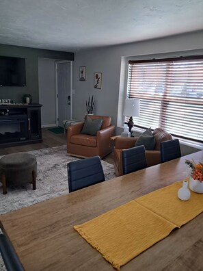 Una sala de estar con un sofá de cuero marrón, una mesa de comedor de madera con sillas, un televisor y una ventana con persianas.