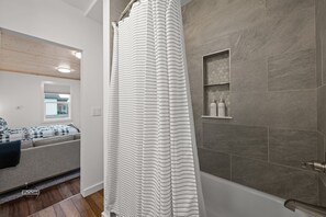 Casa de banho moderna com cortina de chuveiro, prateleira embutida com produtos de higiene e vista para um quarto com sofá.