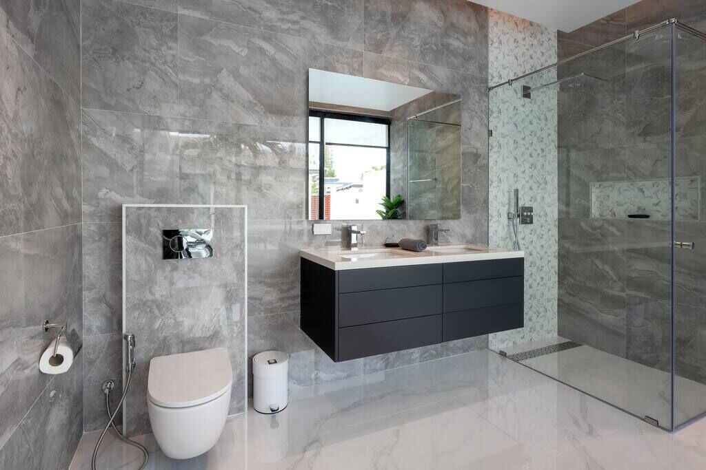 A modern bathroom with a glass shower enclosure, a white toilet, a vanity with a mirror, and marble walls.