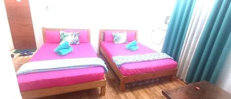 Dos camas de madera con ropa de cama rosa, un taburete de madera y una mesita en una habitación con piso de madera y una puerta de madera.