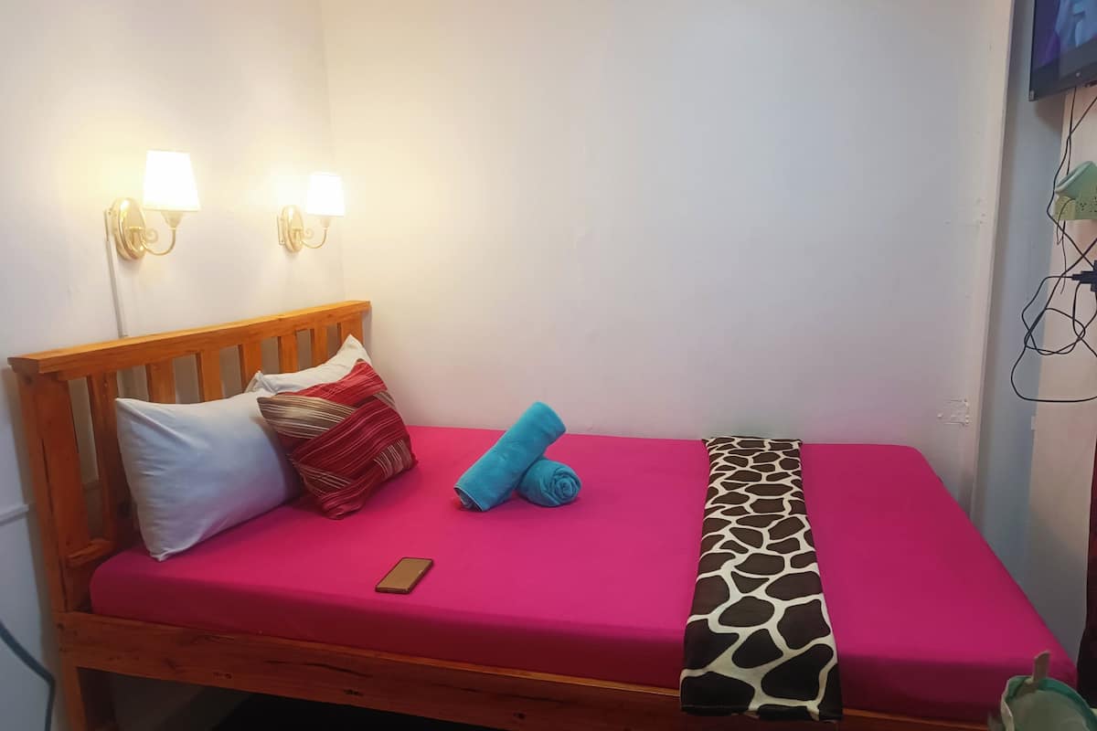 A single bed with a wooden headboard, two wall-mounted lights, a red bedspread, a giraffe-patterned pillow, a blue towel, and a smartphone on the bed.