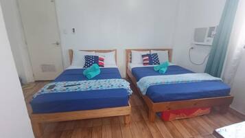 A room with two wooden beds, each with a blue mattress and matching pillows. There is a white door and a small window with a curtain.