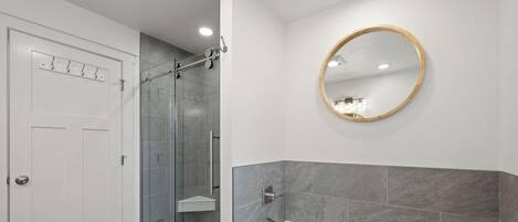 Een badkamer met een douchecabine van glas, een witte deur met haken, een ronde spiegel en een bad met een betegelde muur van marmer.