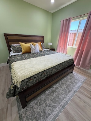 A bedroom with a bed, a nightstand, a lamp, a window with curtains, and a view of the outside. - New Beautiful Gueshouse near Disneyland (Anaheim)