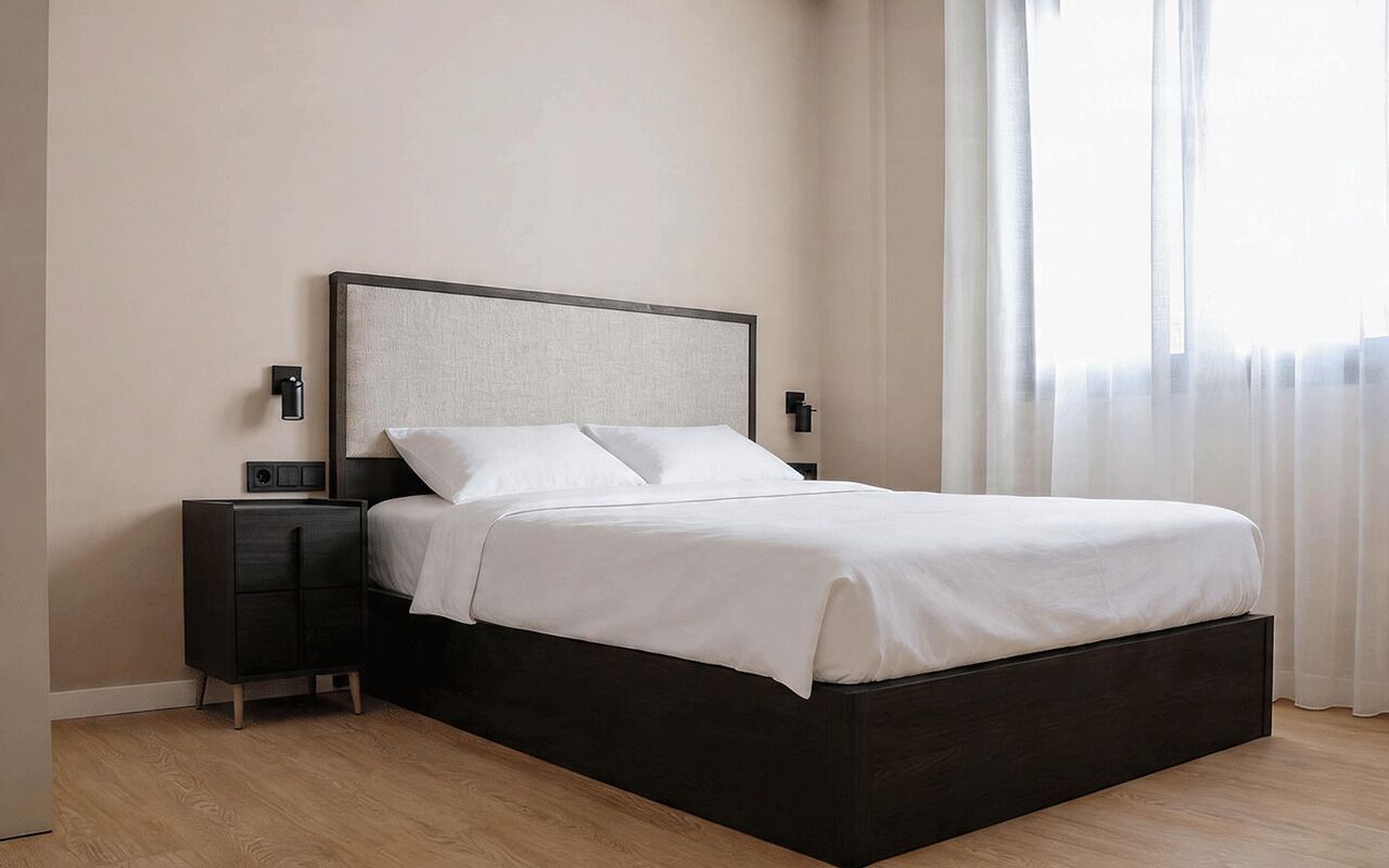 Een bed met donker frame, witte beddengoed, een nachtkastje met een radio en een wandlamp.