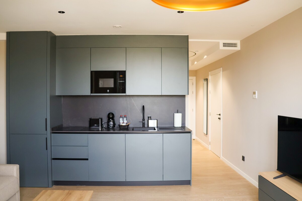 Una cocina moderna con gabinetes grises, microondas integrado y televisor de pantalla plana sobre un pedestal de madera.