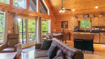 A cabin interior with a wooden ceiling, large windows, a dining area, a kitchen, and a sofa with cushions.