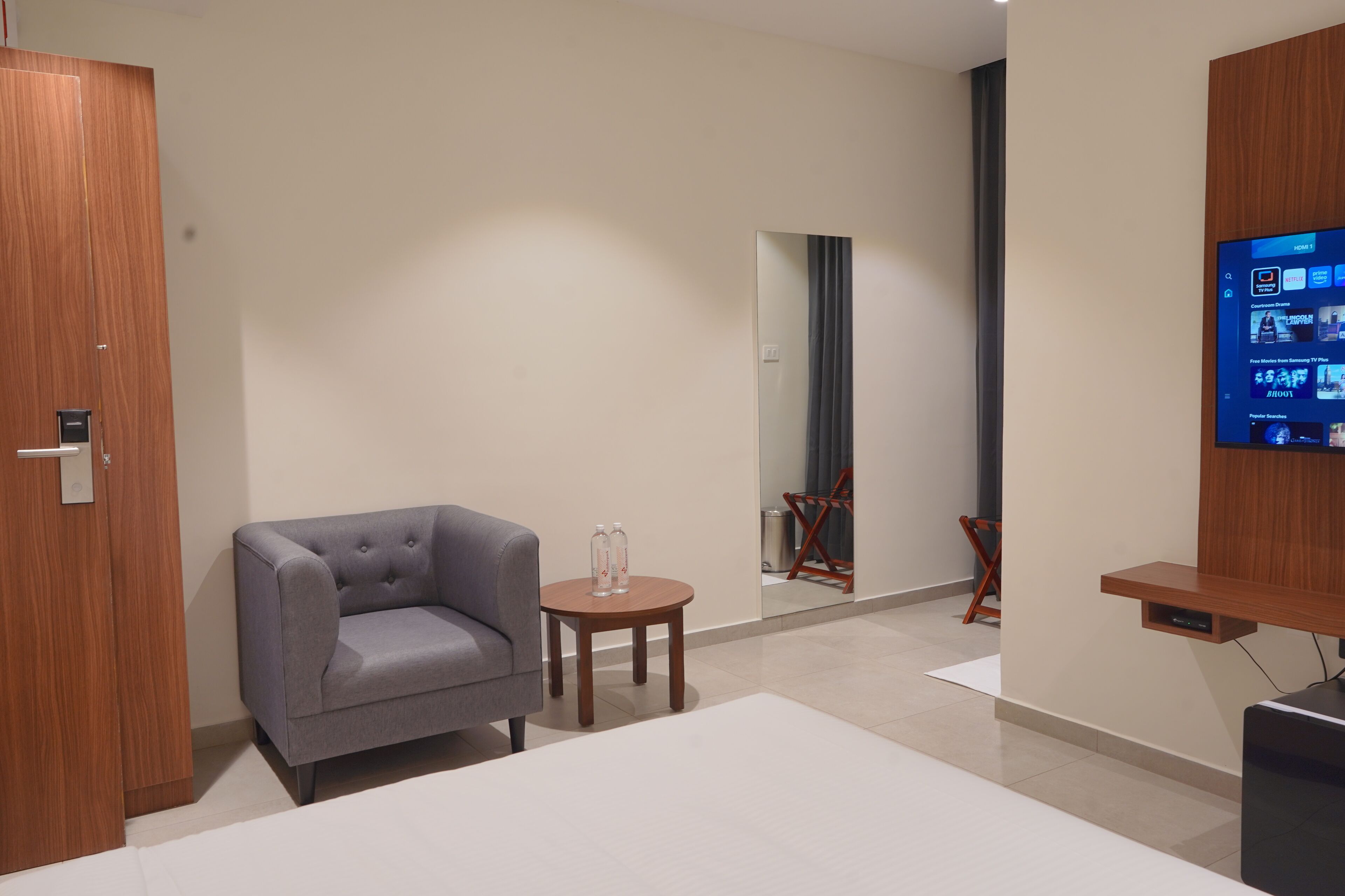 A modern hotel room with a grey sofa, a small round table with two bottles, a TV mounted on the wall, and a wooden wardrobe.