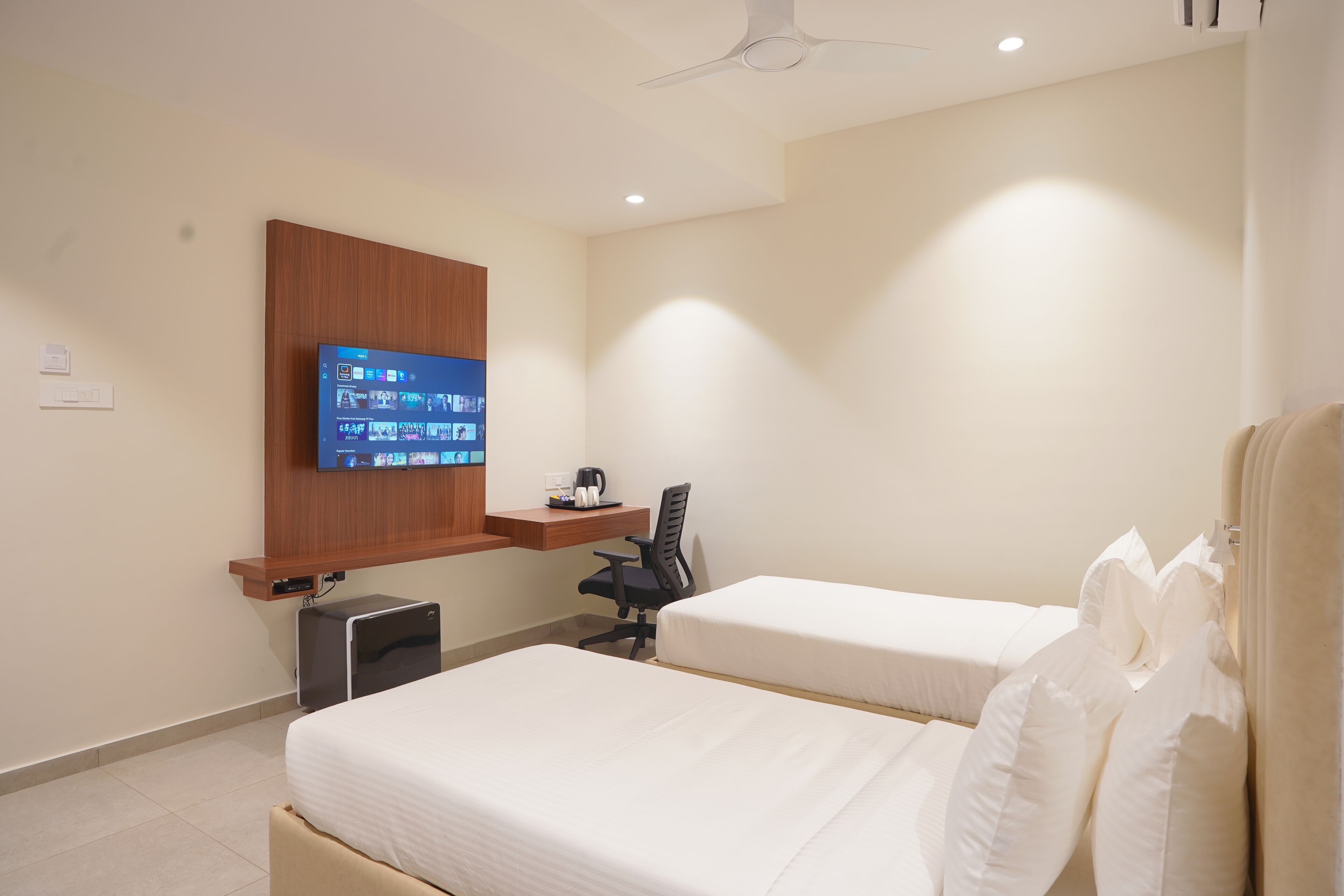 A hotel room with two beds, a desk with a computer, a flat-screen TV mounted on the wall, and a ceiling fan.