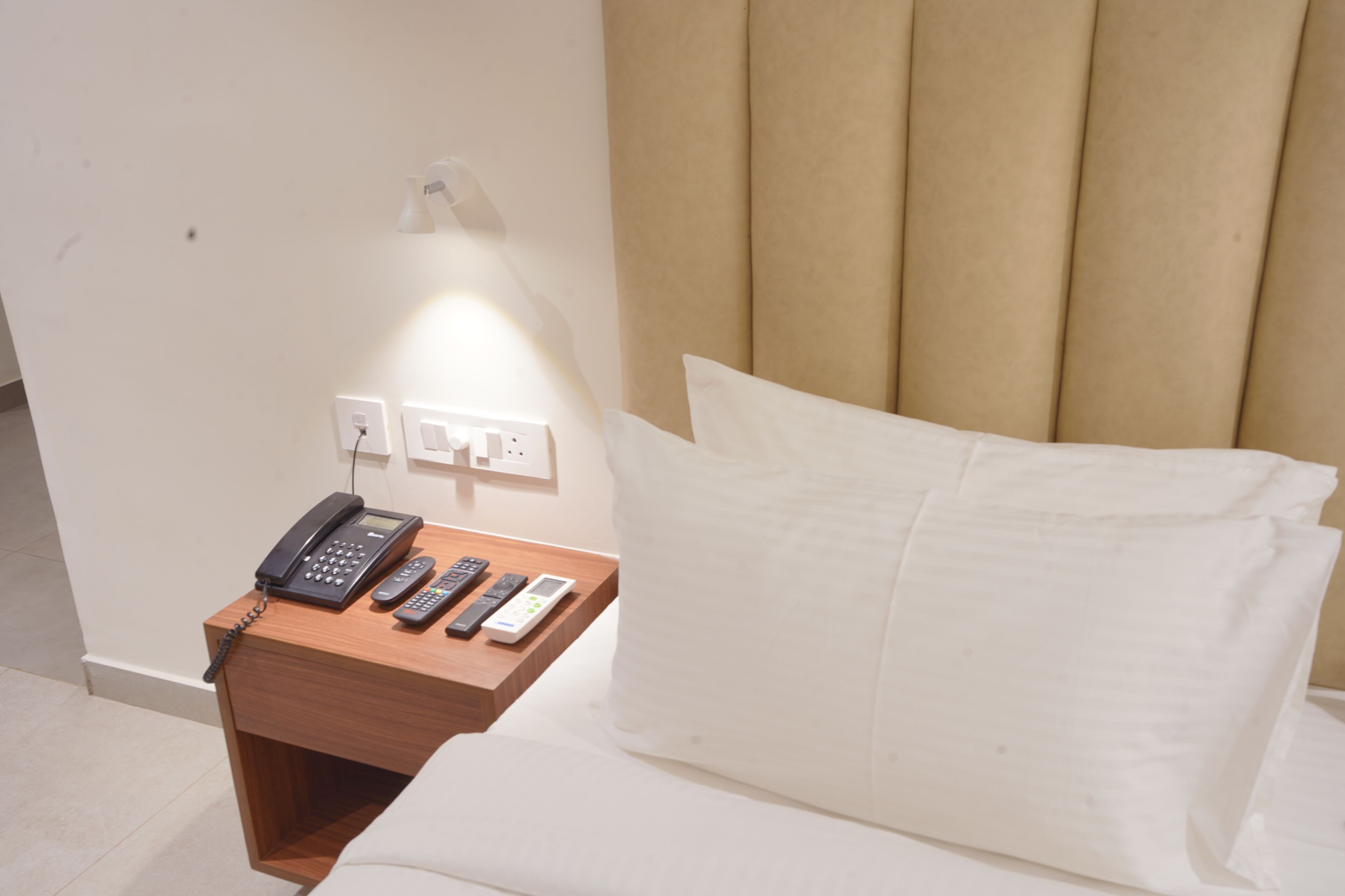 A hotel room with a bed, a wooden nightstand with a phone, remote controls, and a lamp.