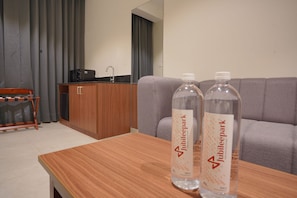 Una habitación con una mesa de madera, dos botellas de agua Jubilee Park, un sofá gris y un mueble de madera con un objeto negro encima.