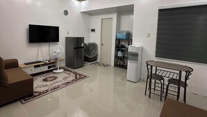 Een moderne woonkamer met een flatscreen televisie, een bank, een eettafel met stoelen, een koelkast, een waterdispenser en een plafondlamp.