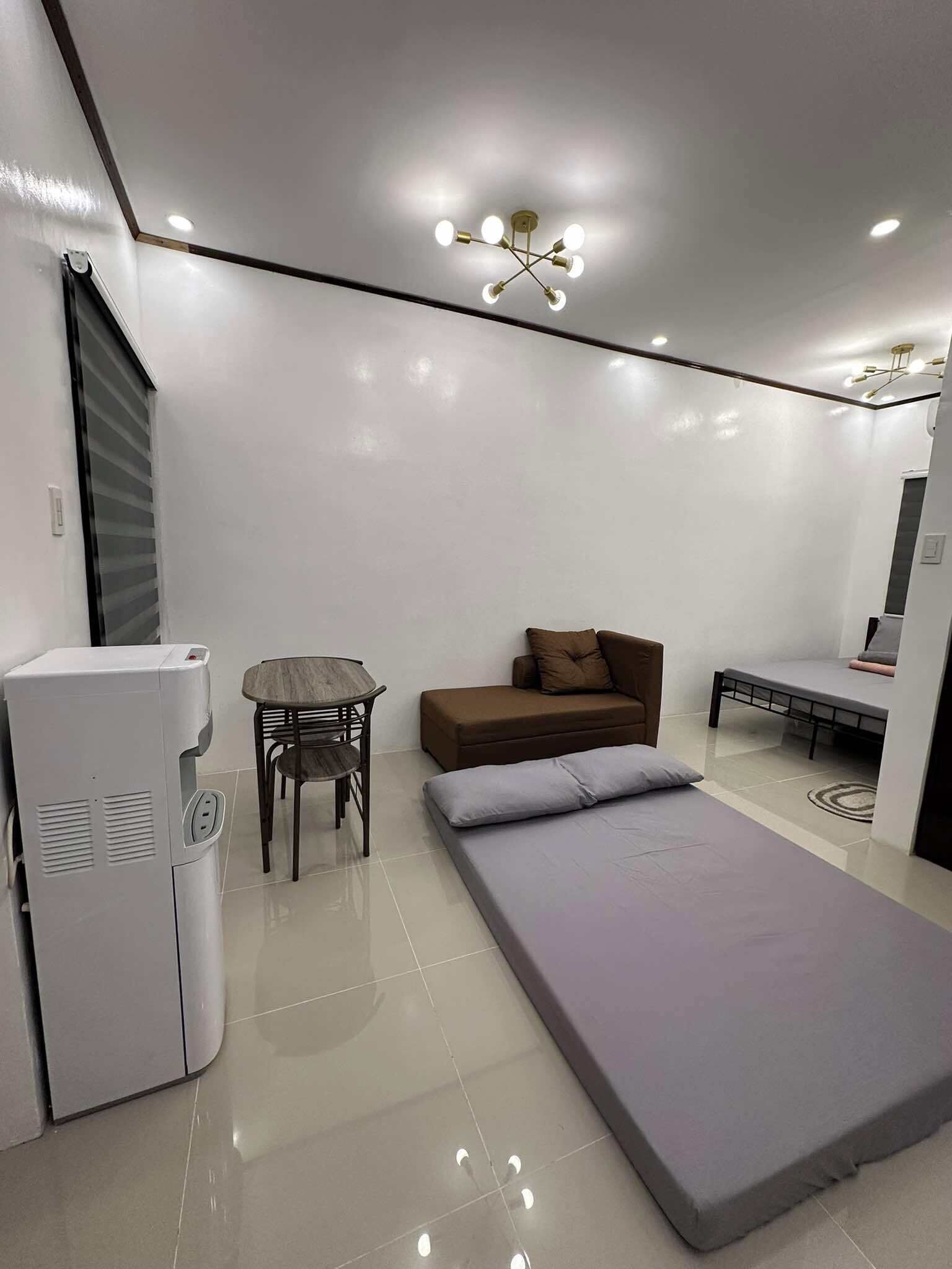 A modern room with a bed, a sofa, a table, and a refrigerator.