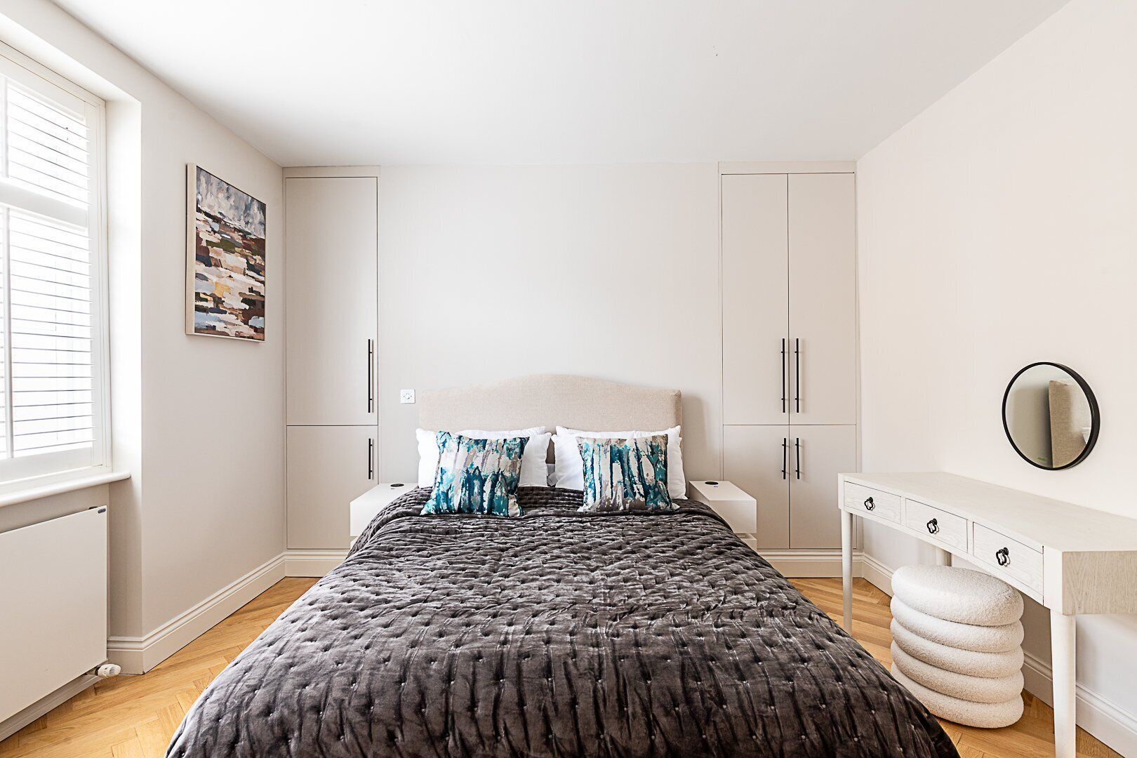A bedroom with a bed, white cabinets, a dresser, a round mirror, and a window with blinds.