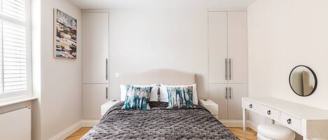 A bedroom with a bed, white cabinets, a dresser, a round mirror, and a window with blinds.