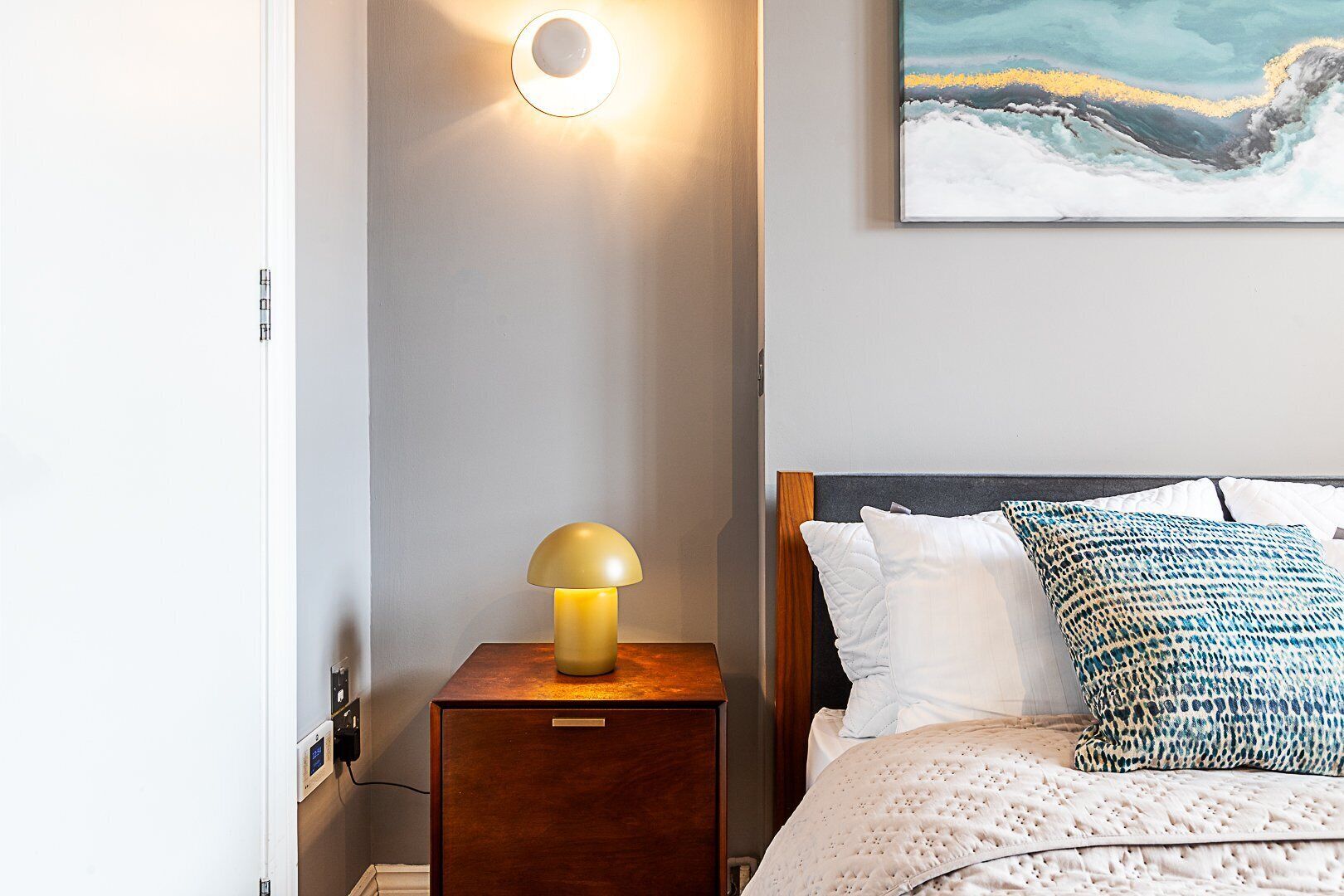 Une chambre à coucher avec un lit, une table de chevet avec une lampe, un luminaire fixé au mur et un tableau.