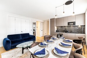 Ein modernes Wohnzimmer mit einem blauen Sofa, einem Esstisch für sechs Personen und einer Küche mit schwarz-weißer Spritzschutzleiste.