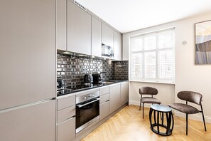 Eine moderne Küche mit grauen Schränken, einer schwarzen, mit Fliesen verkleideten Rückwand, Edelstahlgeräten und einem Holzboden im Fischgrätmuster. Ein Fenster mit Jalousien, ein Stuhl und ein kleiner Tisch sind ebenfalls vorhanden.