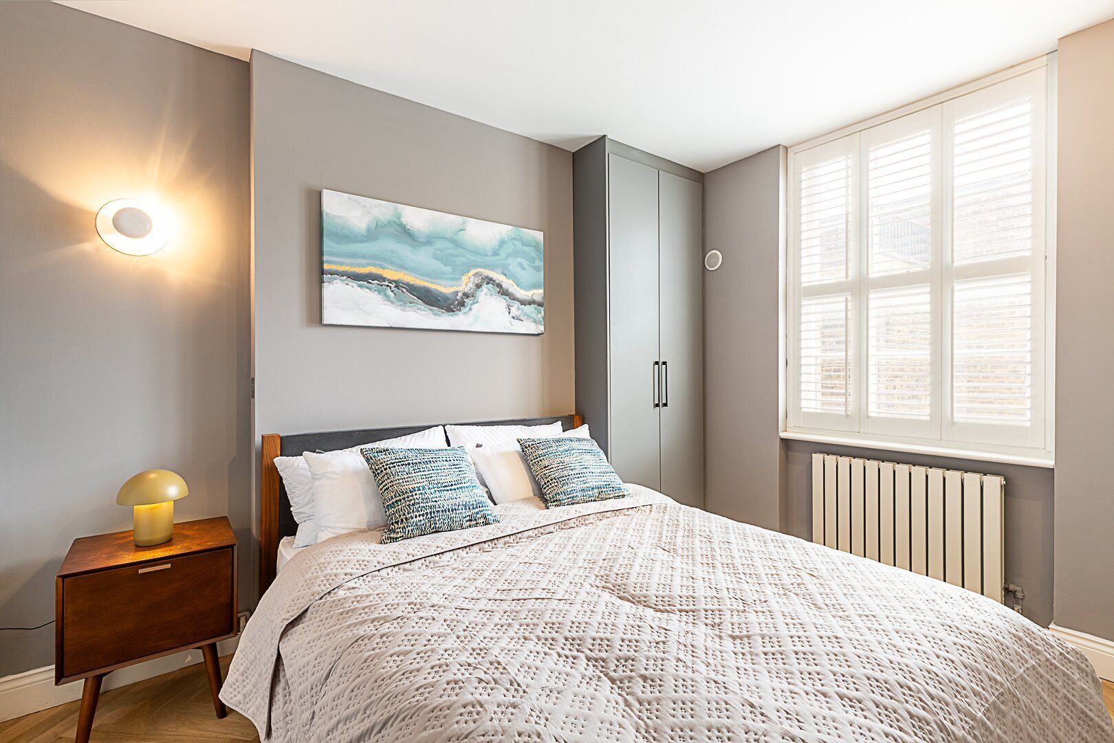 Ein Schlafzimmer mit einem Bett, einem Nachttisch, einer Wandlampe, einem Fenster mit Jalousien und einem Bild an der Wand.
