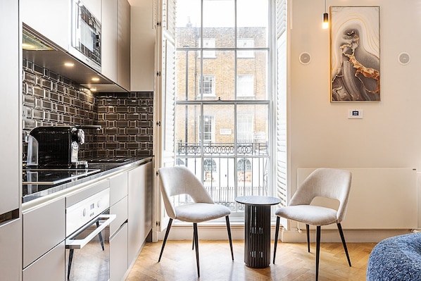 A modern kitchen with a black tiled backsplash, a coffee machine, and a window with a view of buildings. There are two chairs and a small table in the room. - Stylish Marylebone/Baker St Retreat (London)