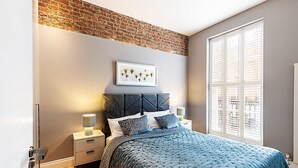 Un dormitorio con pared de ladrillo, cama con colcha azul, mesita de noche con lámpara y ventana con persianas.