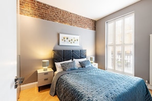 A bedroom with a brick wall, a bed with a blue quilt, a nightstand with a lamp, and a window with blinds. - Stylish Marylebone/Baker St Retreat (London)