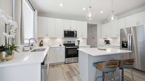Una cocina moderna con armarios blancos, electrodomésticos de acero inoxidable, una isla central con taburetes altos e iluminación colgante.