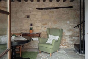A stone-walled room with a green armchair, a small table with a potted plant, and a fireplace.