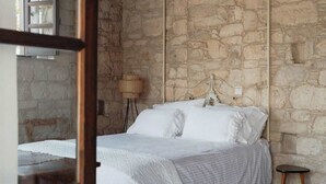 Un dormitorio con pared de piedra, cama con ropa de cama blanca, un taburete pequeño y una lámpara.