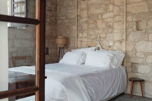 A bedroom with a stone wall, a bed with white bedding, a small stool, and a lamp.