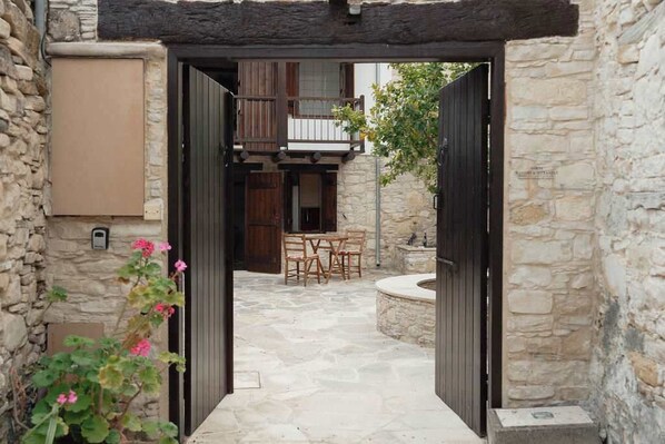 Une cour pavée de pierres, avec une table et des chaises en bois, un mur de pierre et une porte donnant sur un balcon.