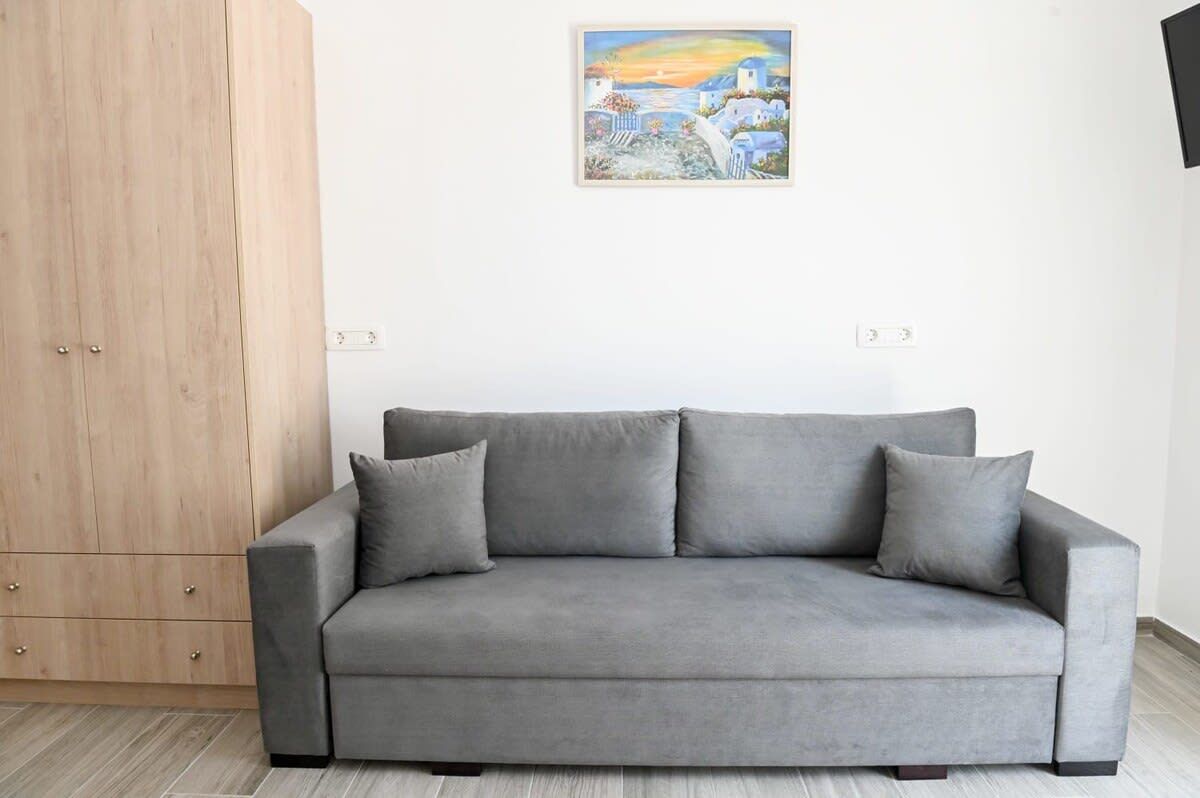 Un sofá gris con tres cojines, un armario de madera y un cuadro con una escena costera en la pared.