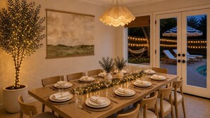 A dining area with a long wooden table set for six, surrounded by chairs, a potted tree with lights, a hanging lamp, and a view of an outdoor pool area. - Pool, Hot Tub, Huge Outdoor Theater, Mini Golf, Bowling, BBQ, Fire Pit,Sleeps 19 (Pflugerville)