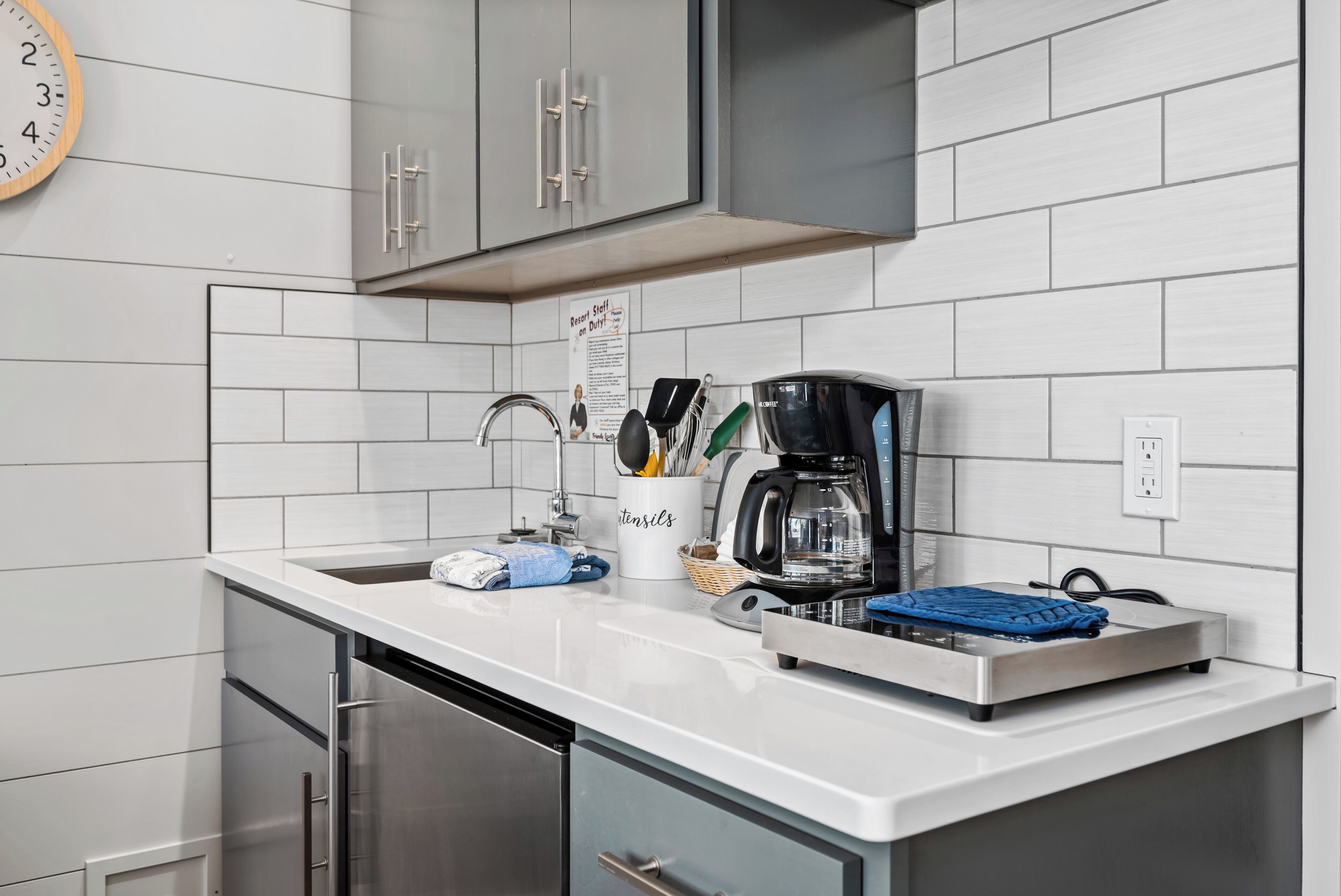 Una cocina con azulejos blancos, electrodomésticos de acero inoxidable, una cafetera y un recipiente para utensilios.